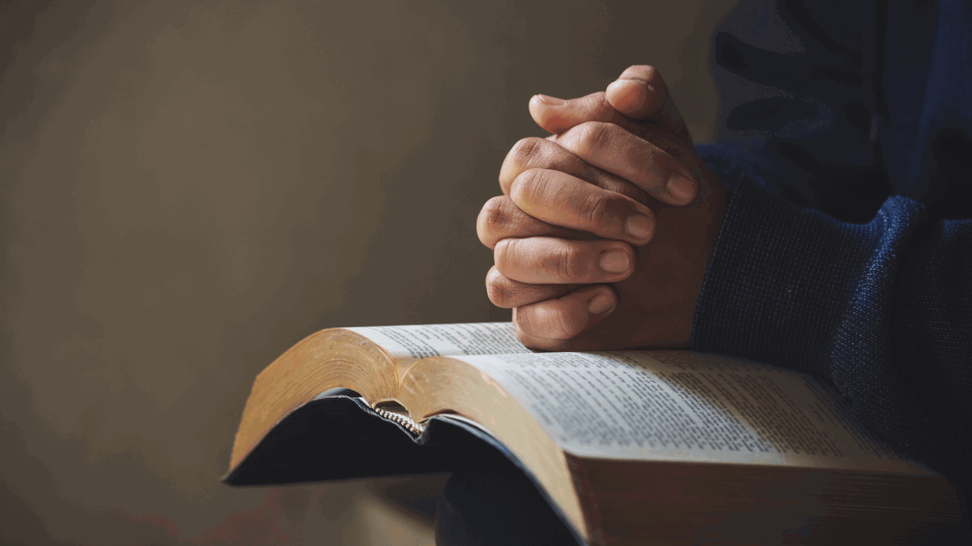 Man's hands in a praying position, resting on a bible.