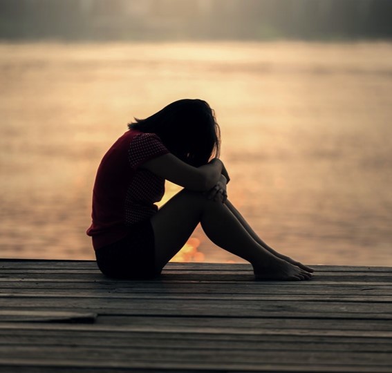 Silhouette of a girl looking sad, resting her head on her knees, by water, at sunset.